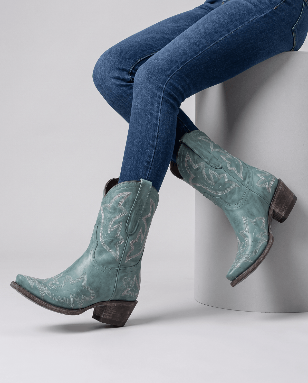 Seated model with blue jeans tucked into teal embroidered boots on a white block