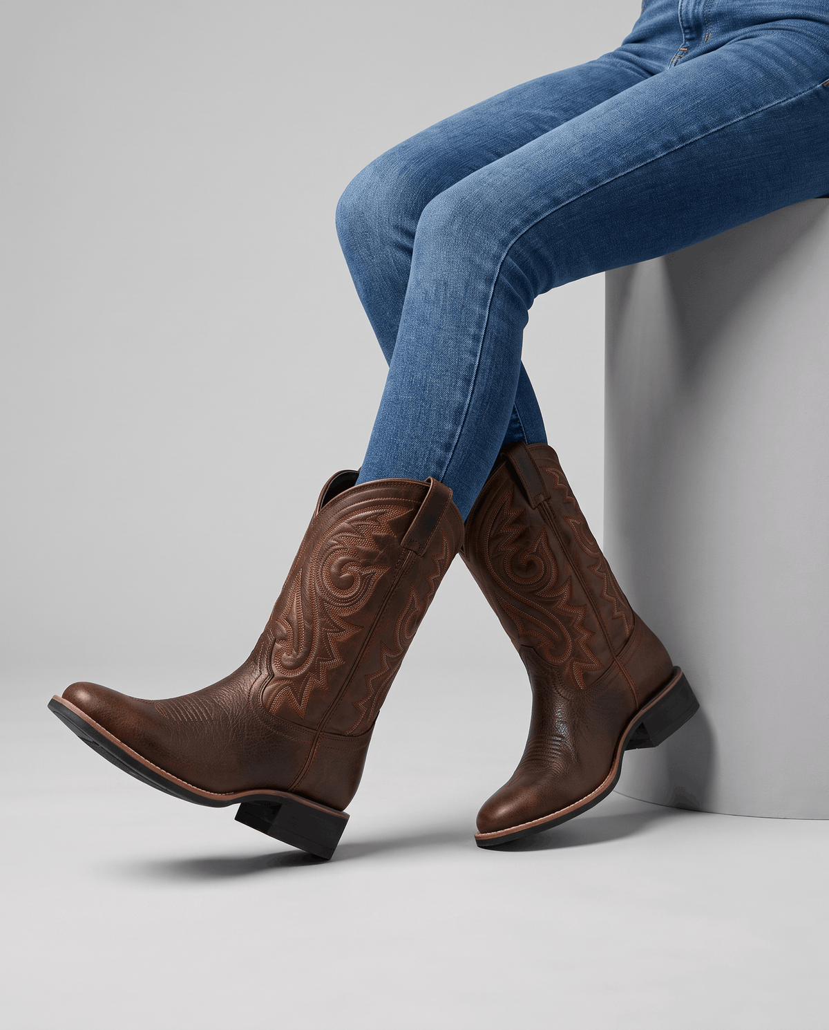 Seated model with blue jeans tucked into brown western boots on a white block