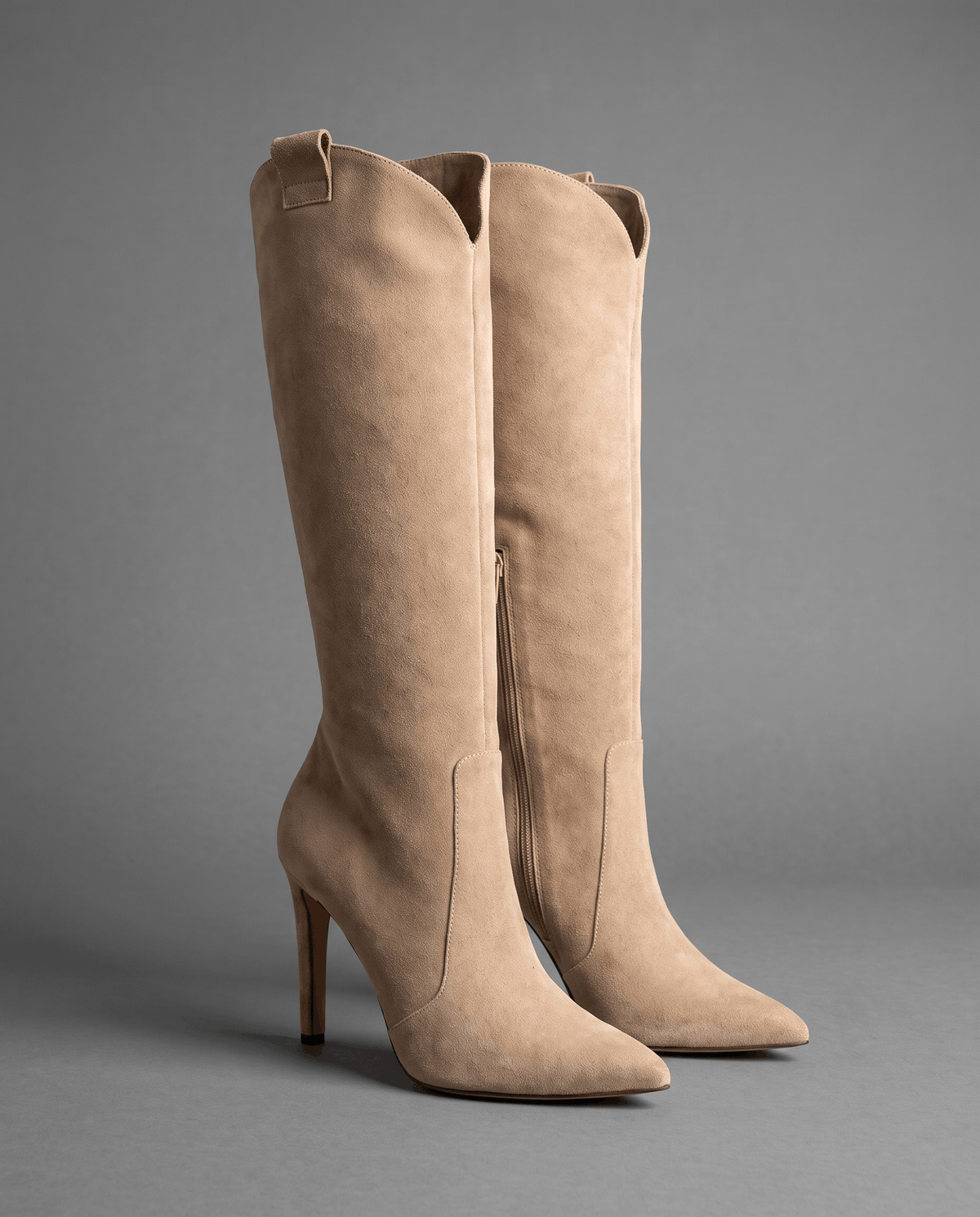 Beige suede knee-high boots with a pointed toe, slim stiletto heel, close-cut shaft, side zip, and collar pull tab.