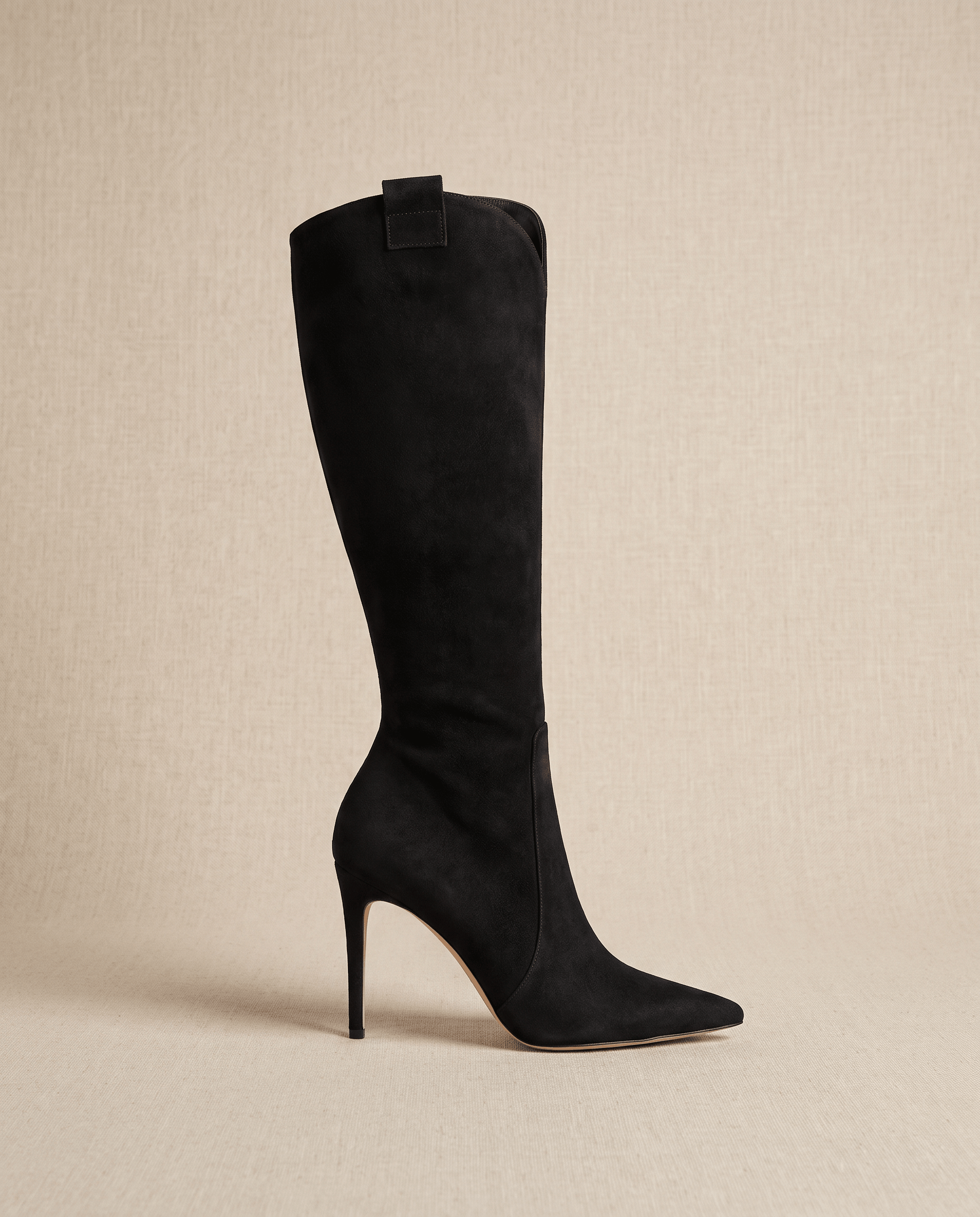 Black suede knee-high boots with a pointed toe, slim stiletto heel, close-cut shaft, and collar pull tab.