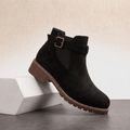 Black suede ankle boots with buckle details and a block heel displayed on a white pedestal.