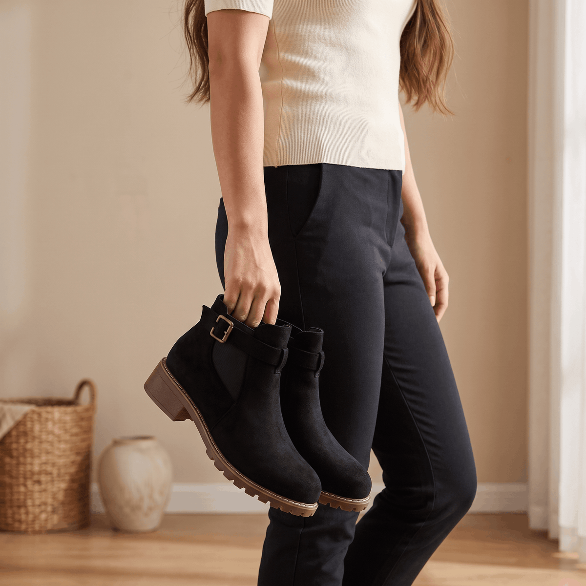 Woman holding black suede ankle boots while wearing dark jeans.