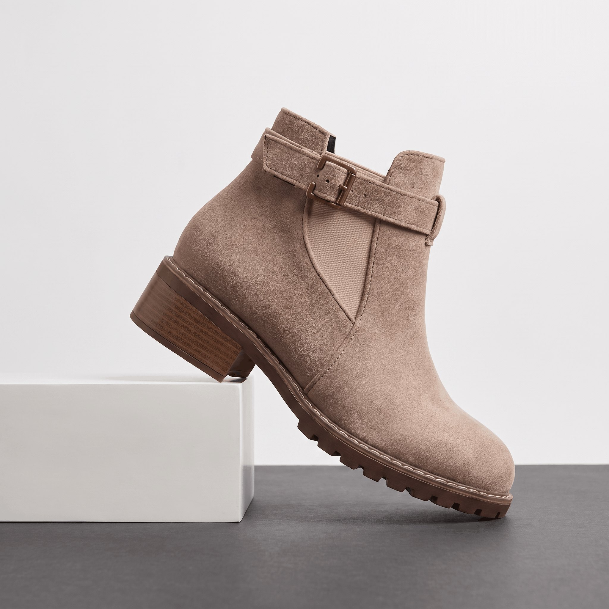 Light beige suede ankle boot with buckle detail and block heel displayed on a white pedestal.