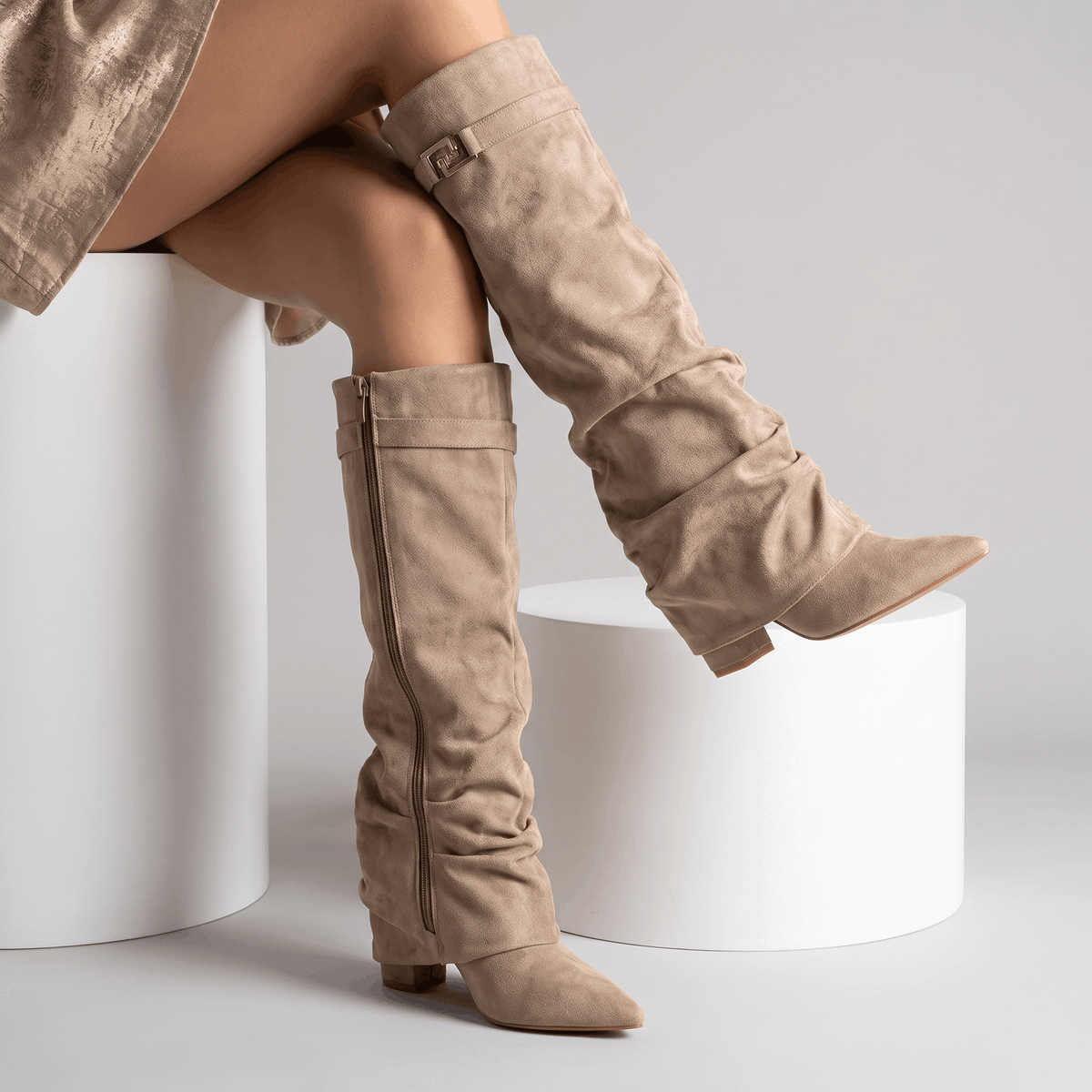Woman seated wearing taupe suede fold-over knee-high boots with a low block heel.