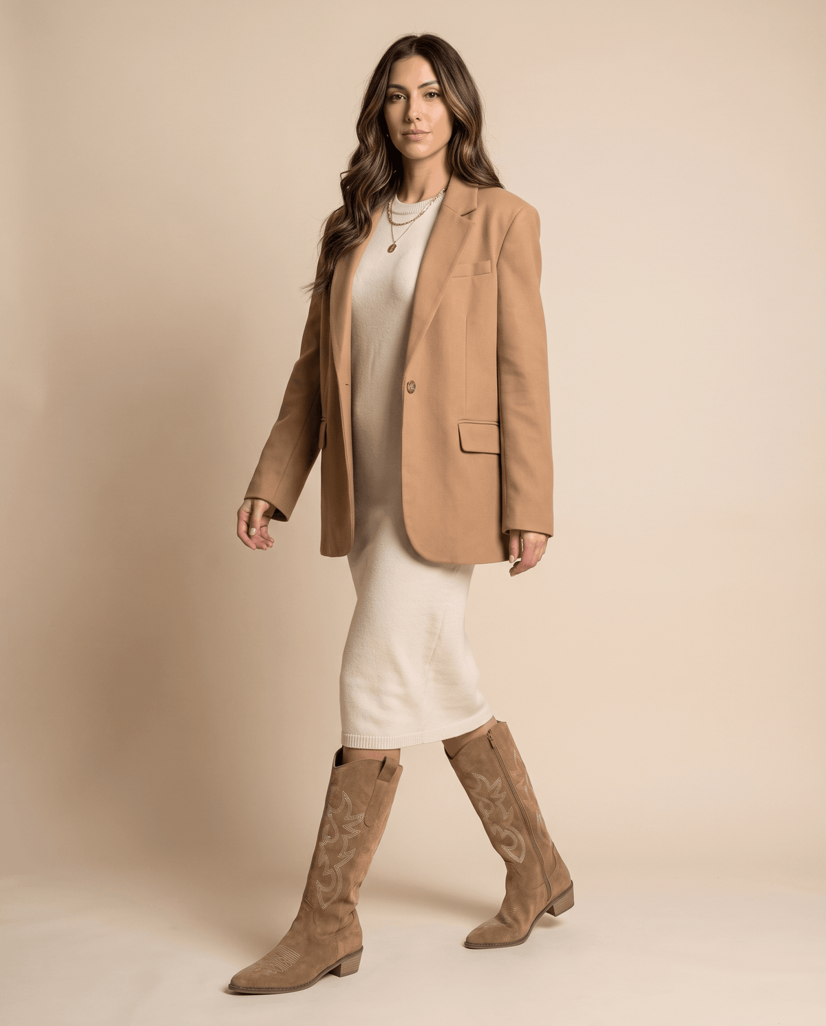 Woman in a camel blazer and cream slip dress walking in brown western boots