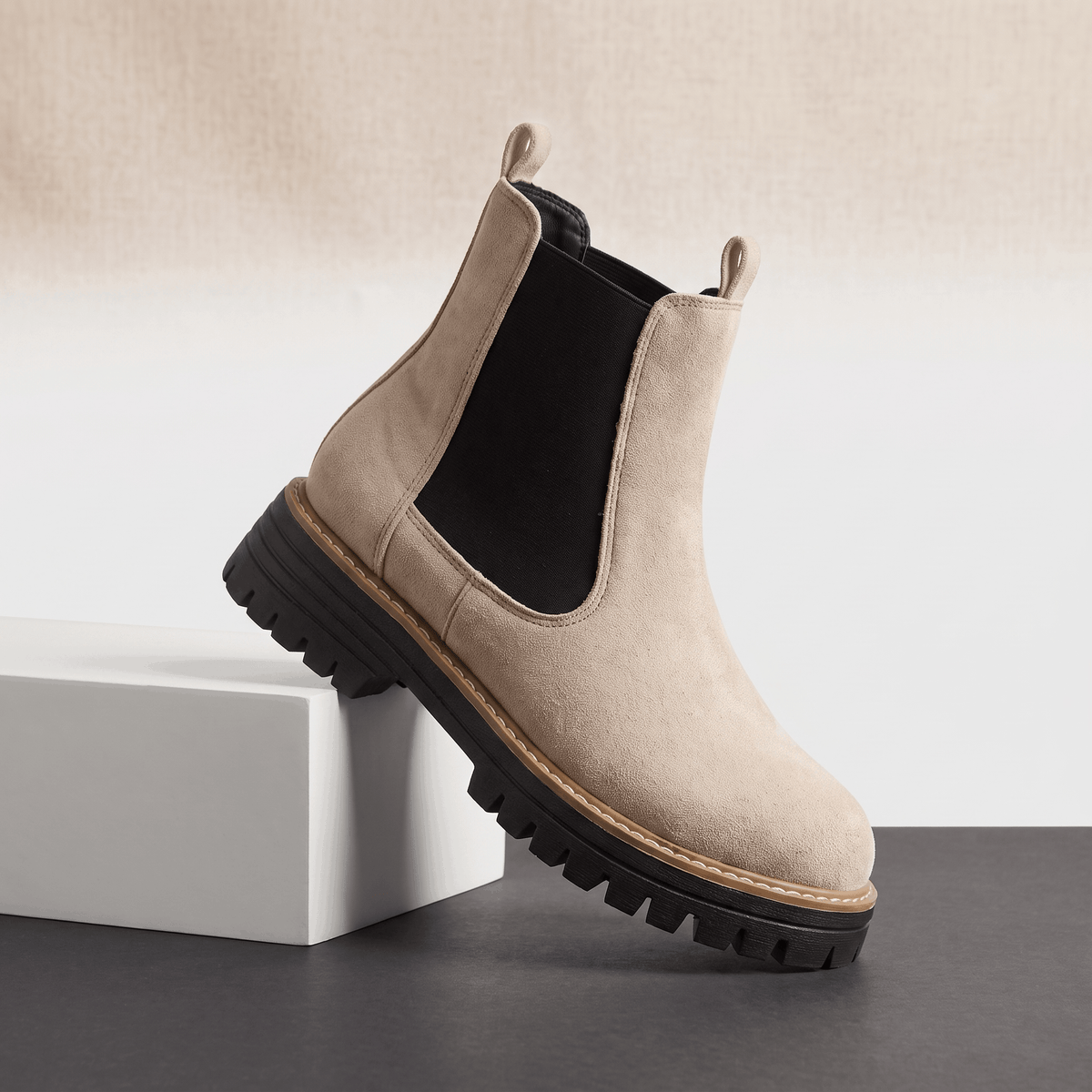 Beige suede Chelsea ankle boot with black elastic panels and lug sole displayed on a white pedestal.