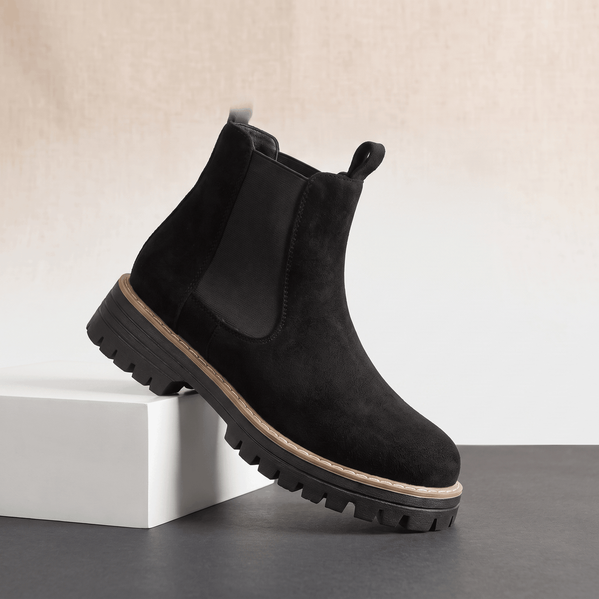 Black suede Chelsea ankle boot with lug sole displayed on a white pedestal.