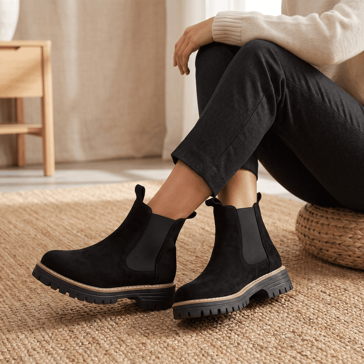 Woman wearing black suede Chelsea ankle boots with black trousers in a cozy indoor setting.