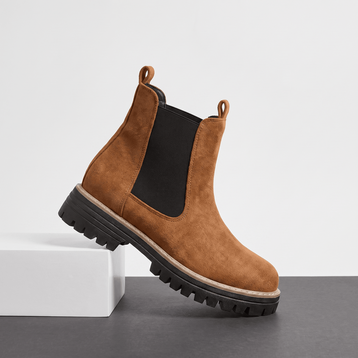Brown suede Chelsea ankle boot with a lug sole displayed on a white pedestal.