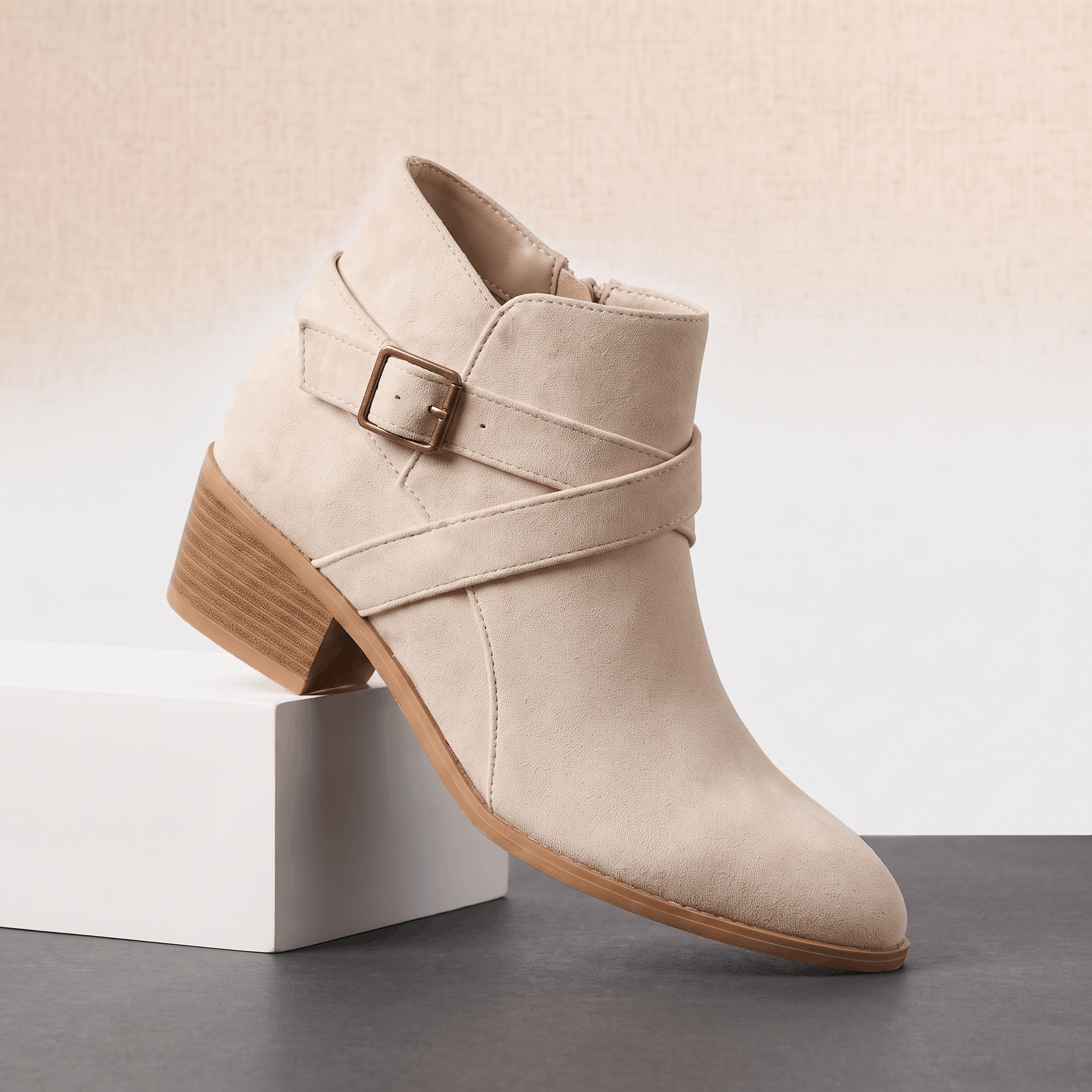Beige suede ankle boot with layered wraparound straps, buckle detail, and a block heel displayed on a white pedestal.