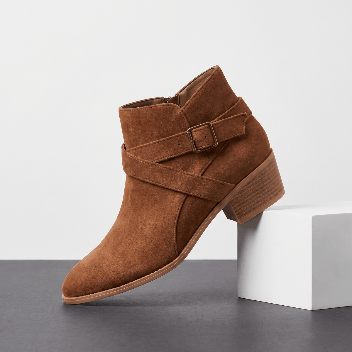 Brown suede ankle boot with side buckle displayed on a white pedestal.