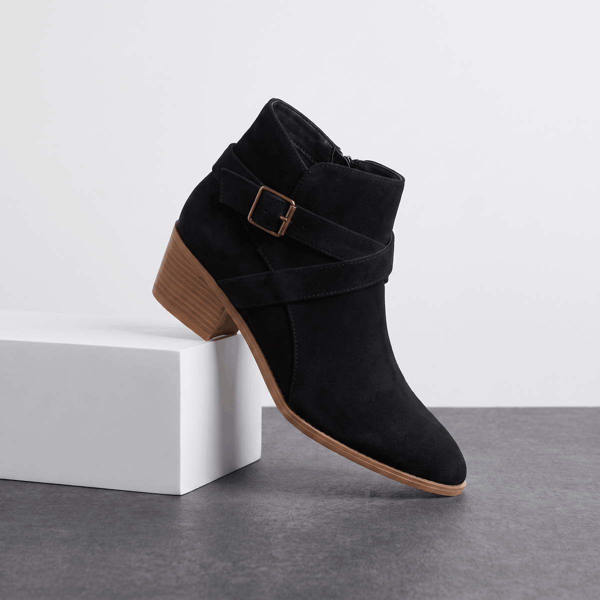 Black suede ankle boot with side buckle displayed on a white pedestal.