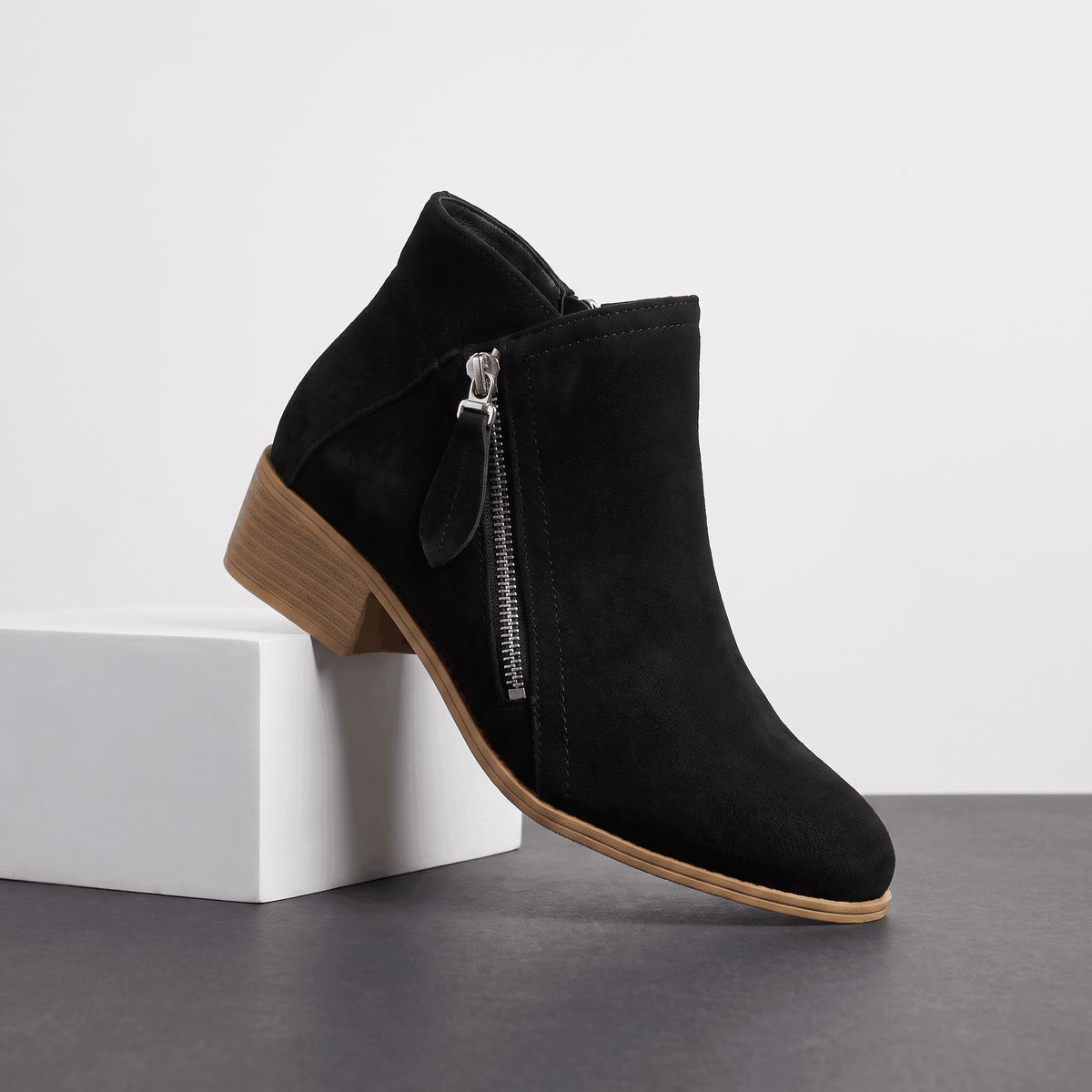 Black suede ankle boot with a side zipper and block heel displayed on a white pedestal.