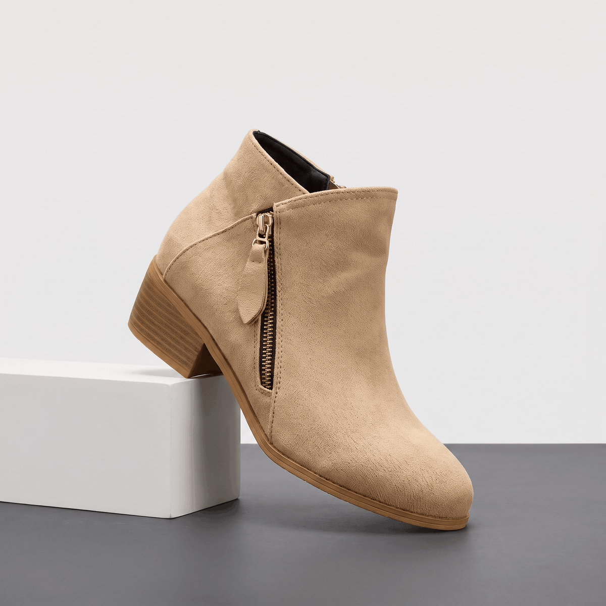 Beige suede ankle boot with a side zipper and block heel displayed on a white pedestal.