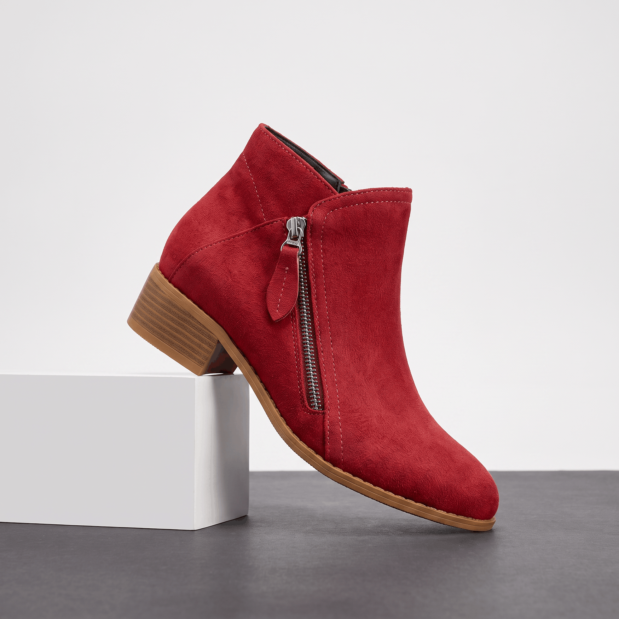 Red suede ankle boot with a side zipper and block heel displayed on a white pedestal.