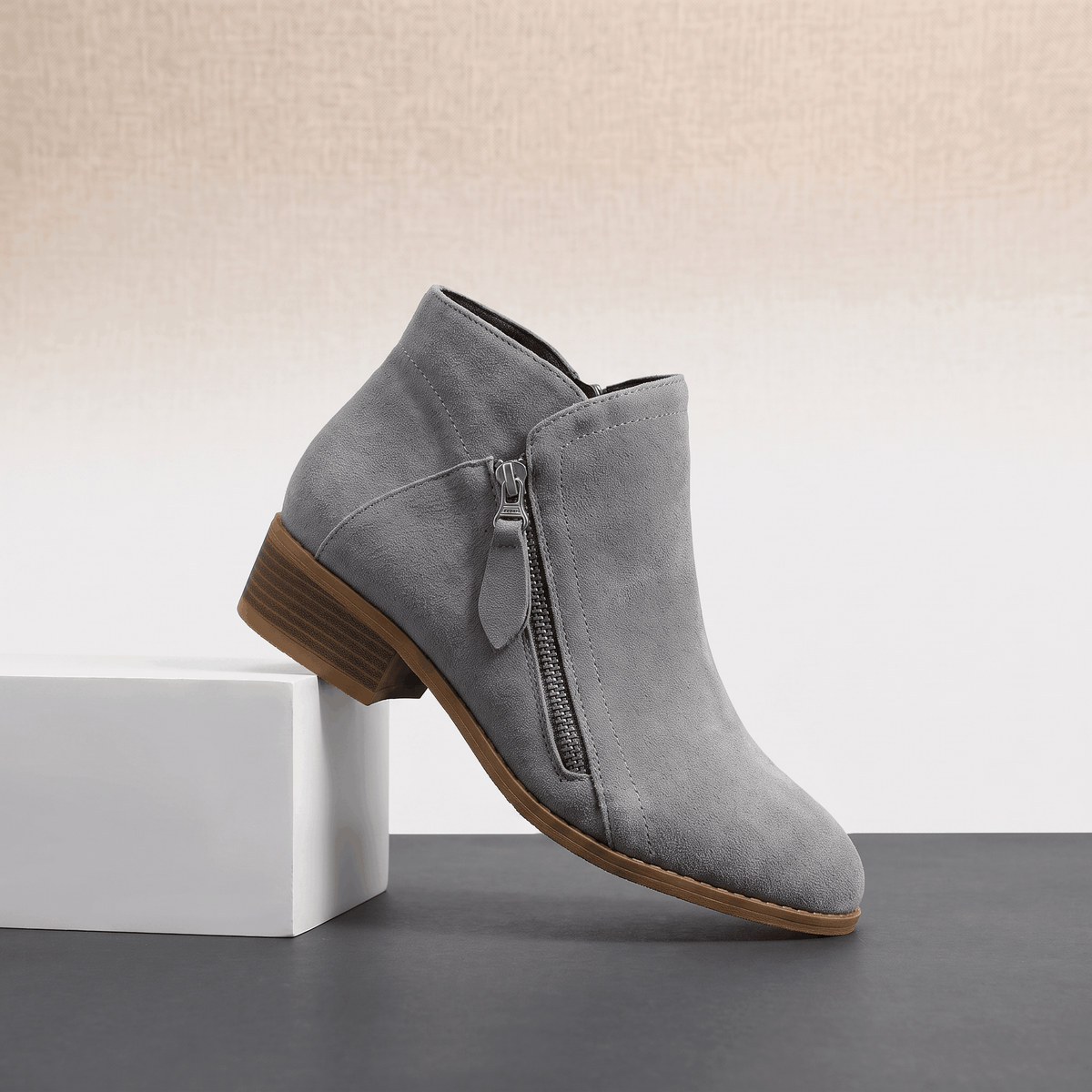 Gray suede ankle boot with a side zipper and block heel displayed on a white pedestal.