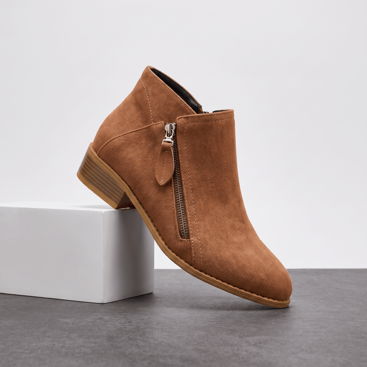 Brown suede ankle boot with a side zipper and block heel displayed on a white pedestal.