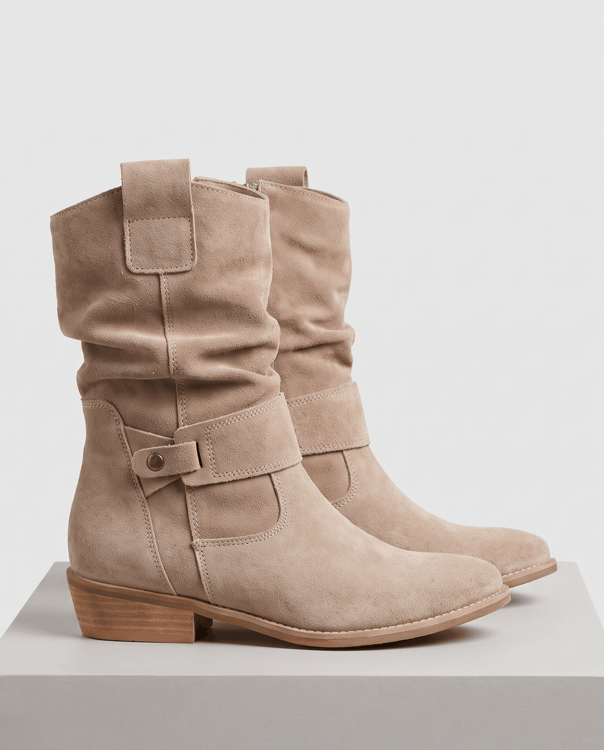 Pair of beige suede slouch boots displayed on a white pedestal