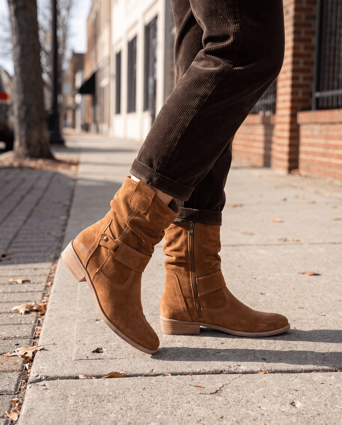 Tan suede slouch boots worn with dark trousers on a sidewalk