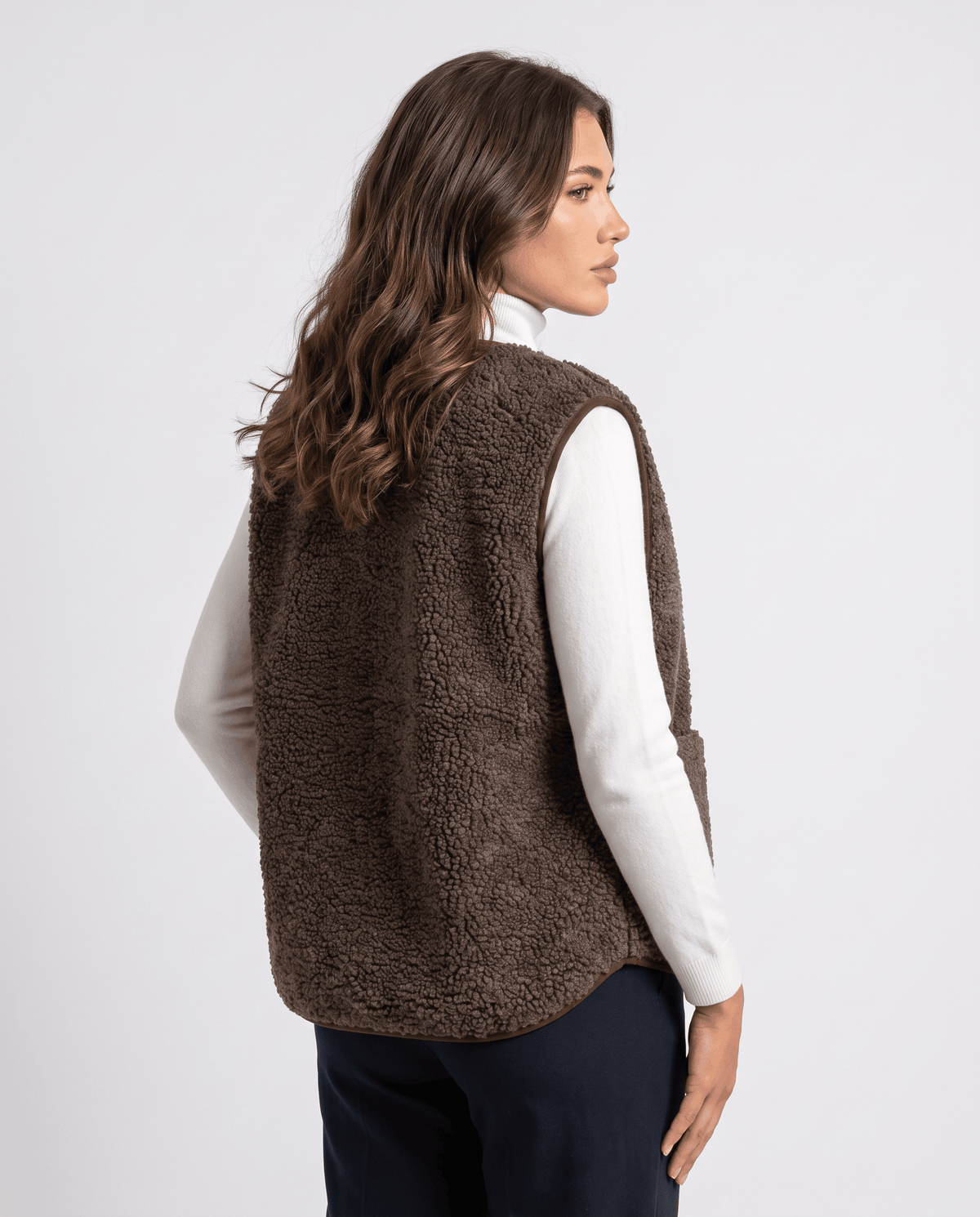 Back three-quarter view of brown teddy fleece vest over a white turtleneck and dark trousers