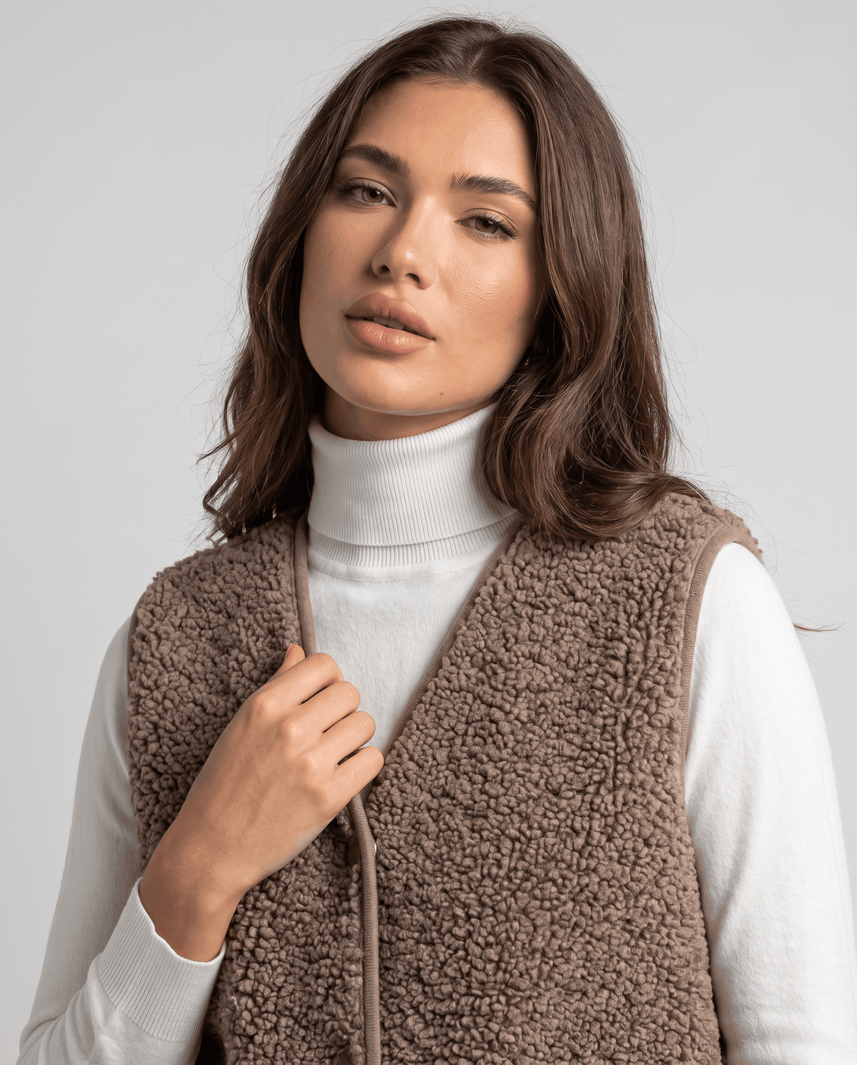 Close-up of taupe teddy fleece vest with one hand at the button front over a turtleneck