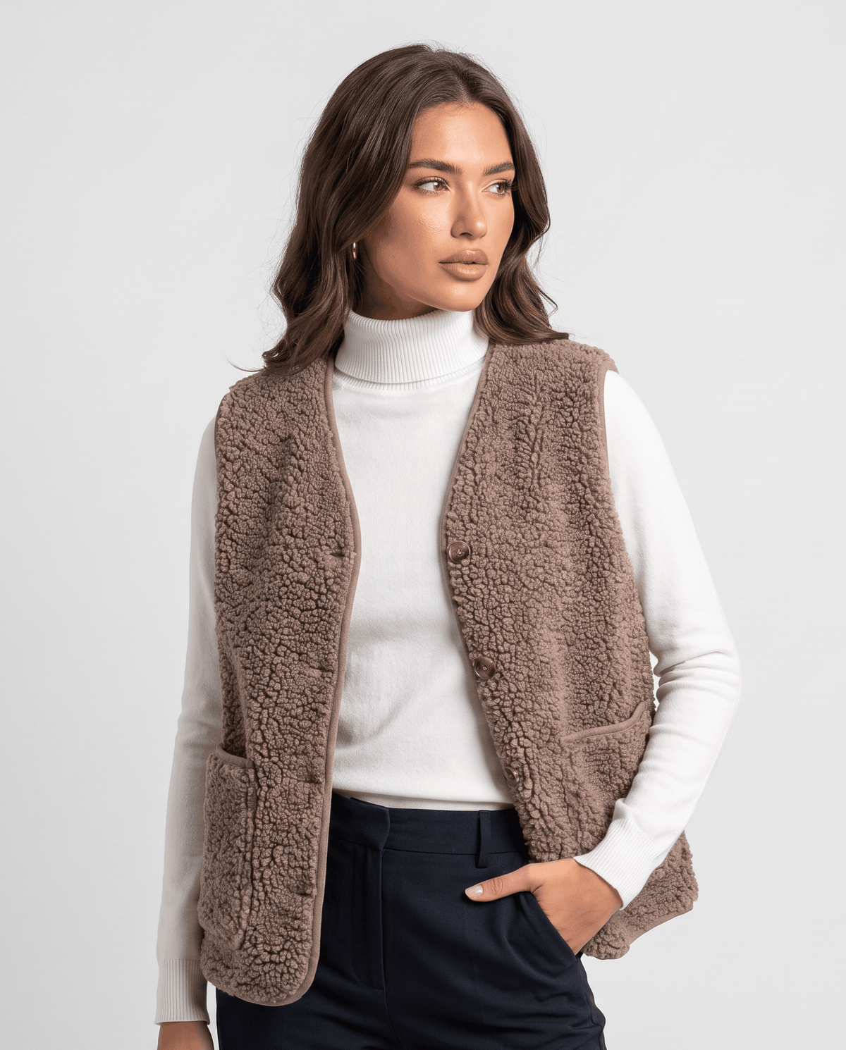 Taupe teddy fleece vest over a white turtleneck, one hand in the front patch pocket