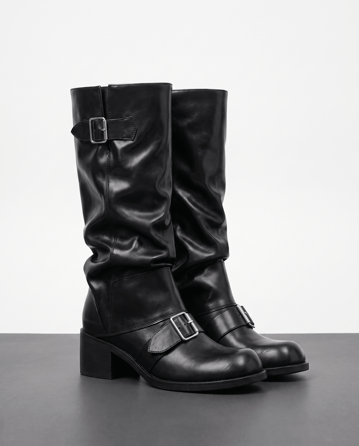 Black vegan leather mid-calf boots with ruched slouch shafts, dual buckle straps, and stacked block heels.
