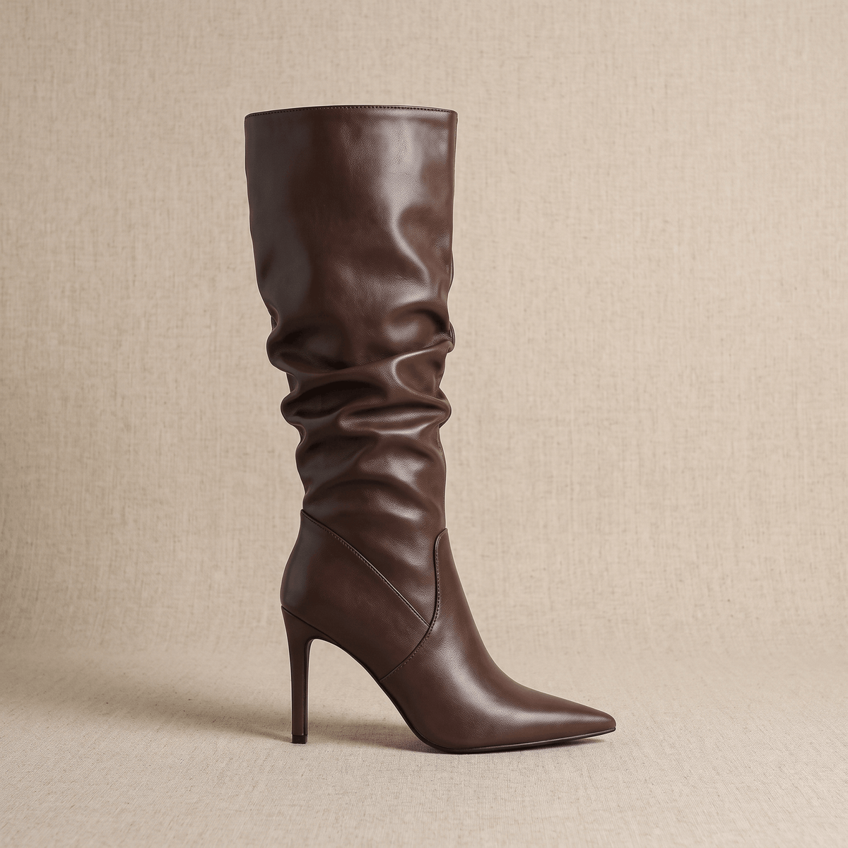 Brown vegan leather knee-high stiletto boots with a pointed toe and ruched, slouchy shaft on a neutral background.