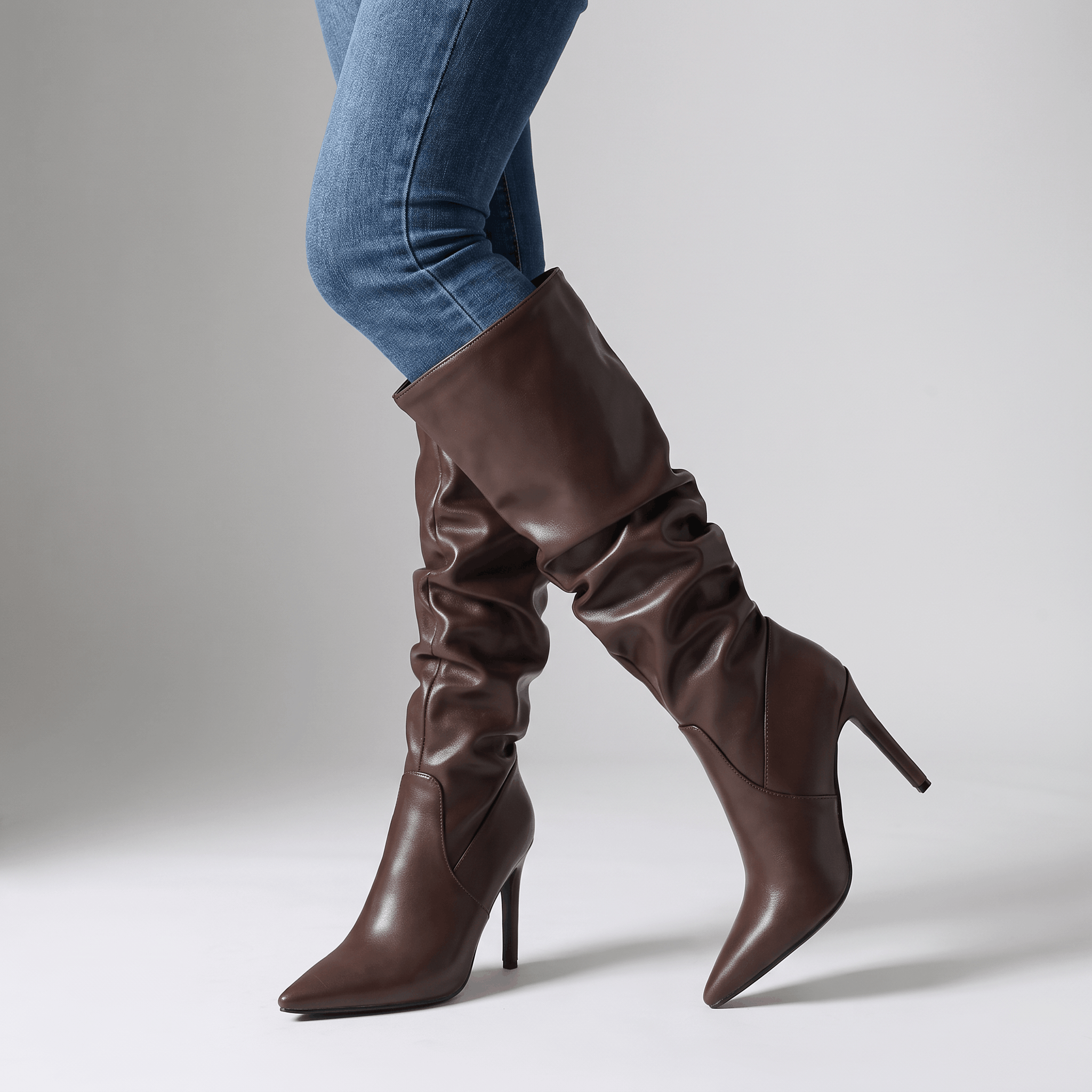 Brown vegan leather knee-high boots with a ruched slouch shaft, pointed toe, and slim stiletto heel worn over blue jeans.