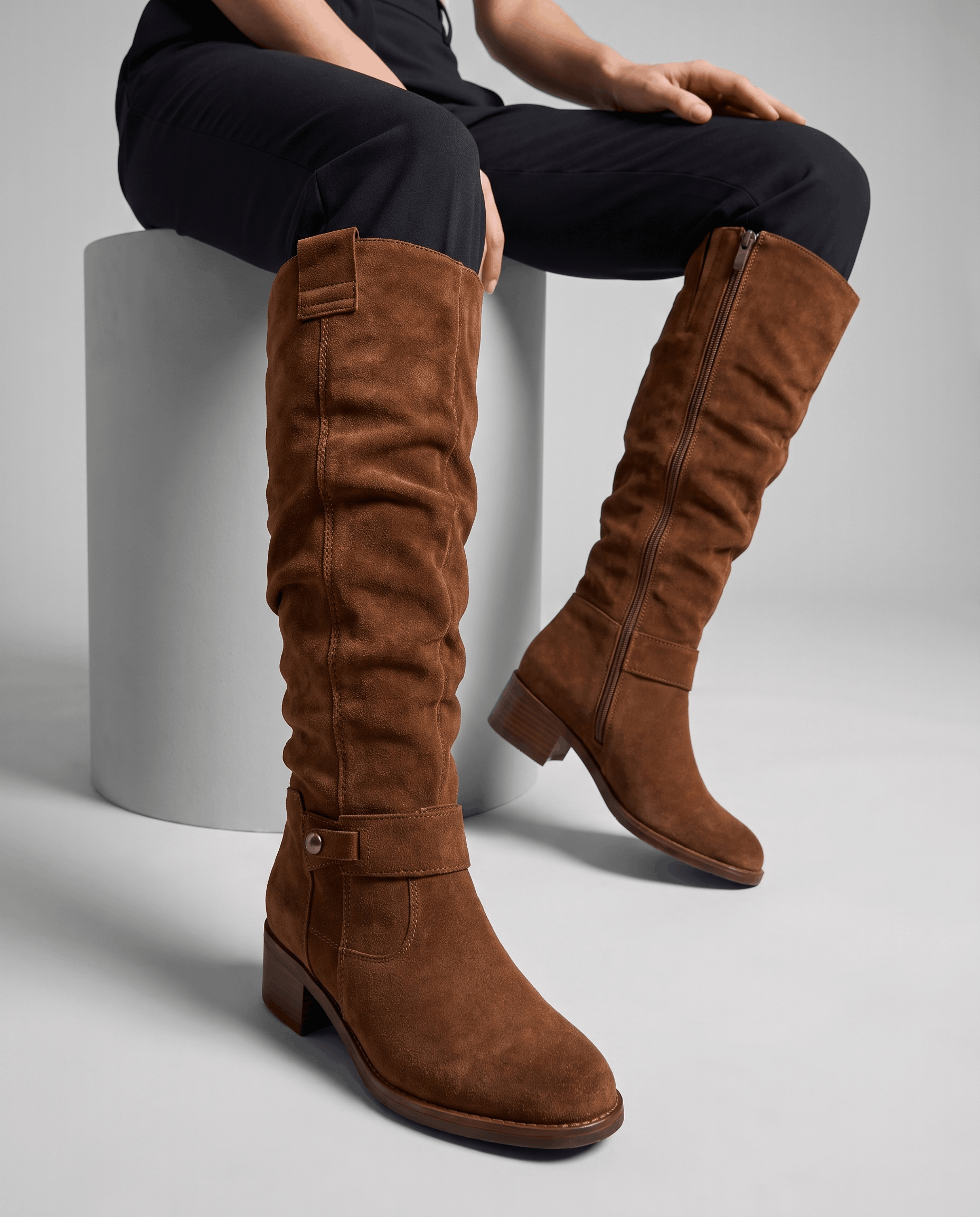 Woman seated wearing tall dark brown vegan suede slouch boots with a low stacked heel.