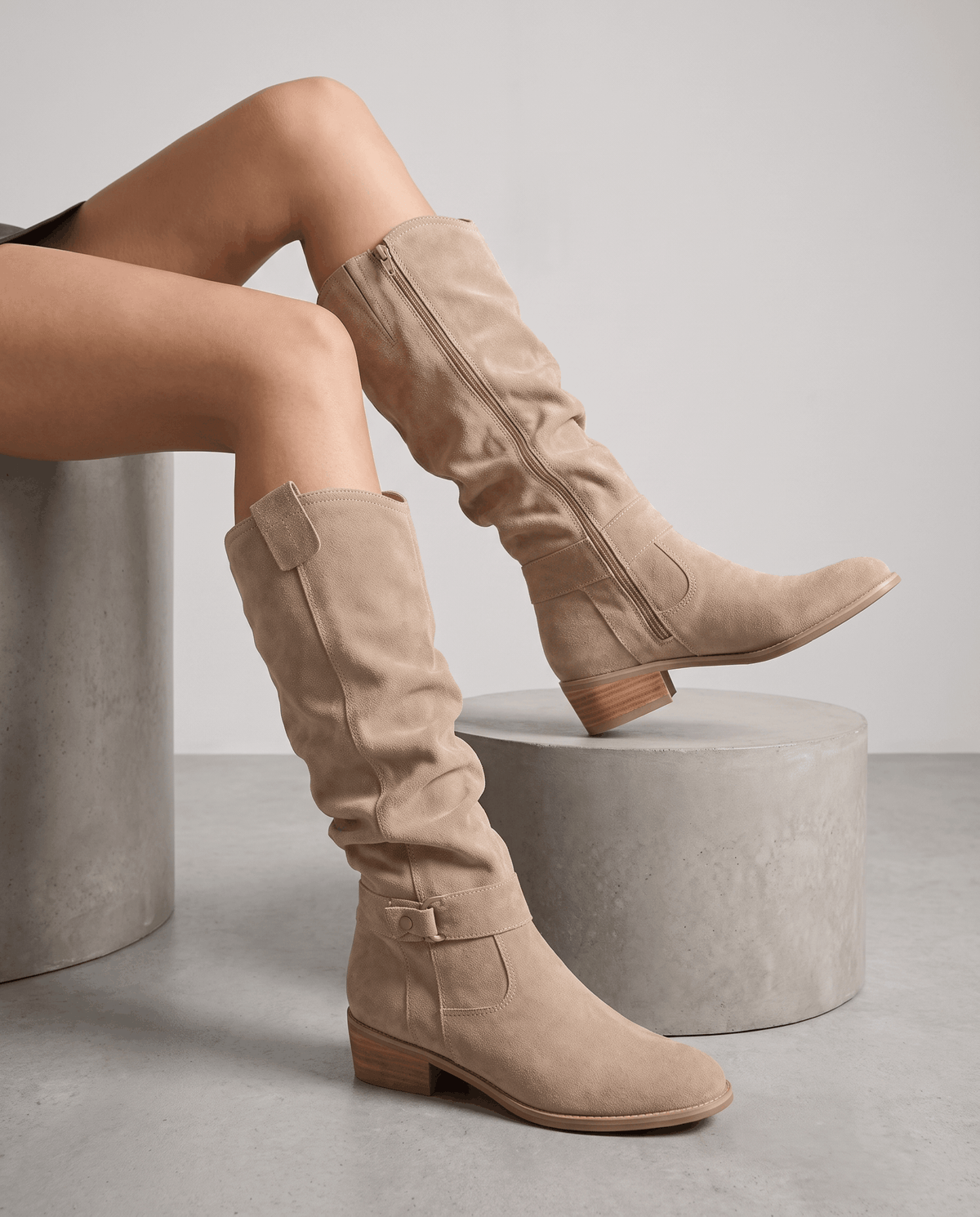 Woman seated on concrete plinths wearing beige vegan suede knee-high boots with a full side zip.