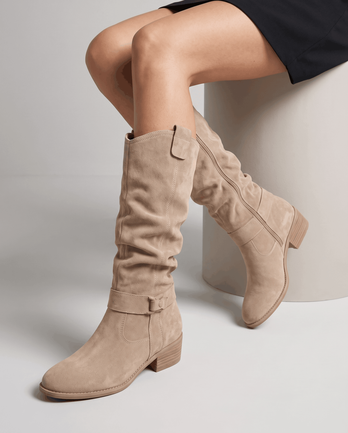 Woman seated wearing beige vegan suede knee-high slouch boots with a black skirt.