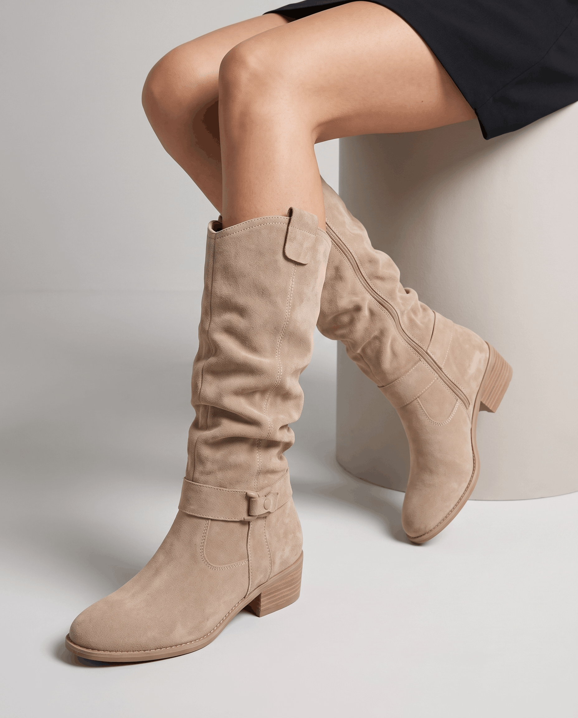 Woman seated wearing beige vegan suede knee-high slouch boots with a black skirt.