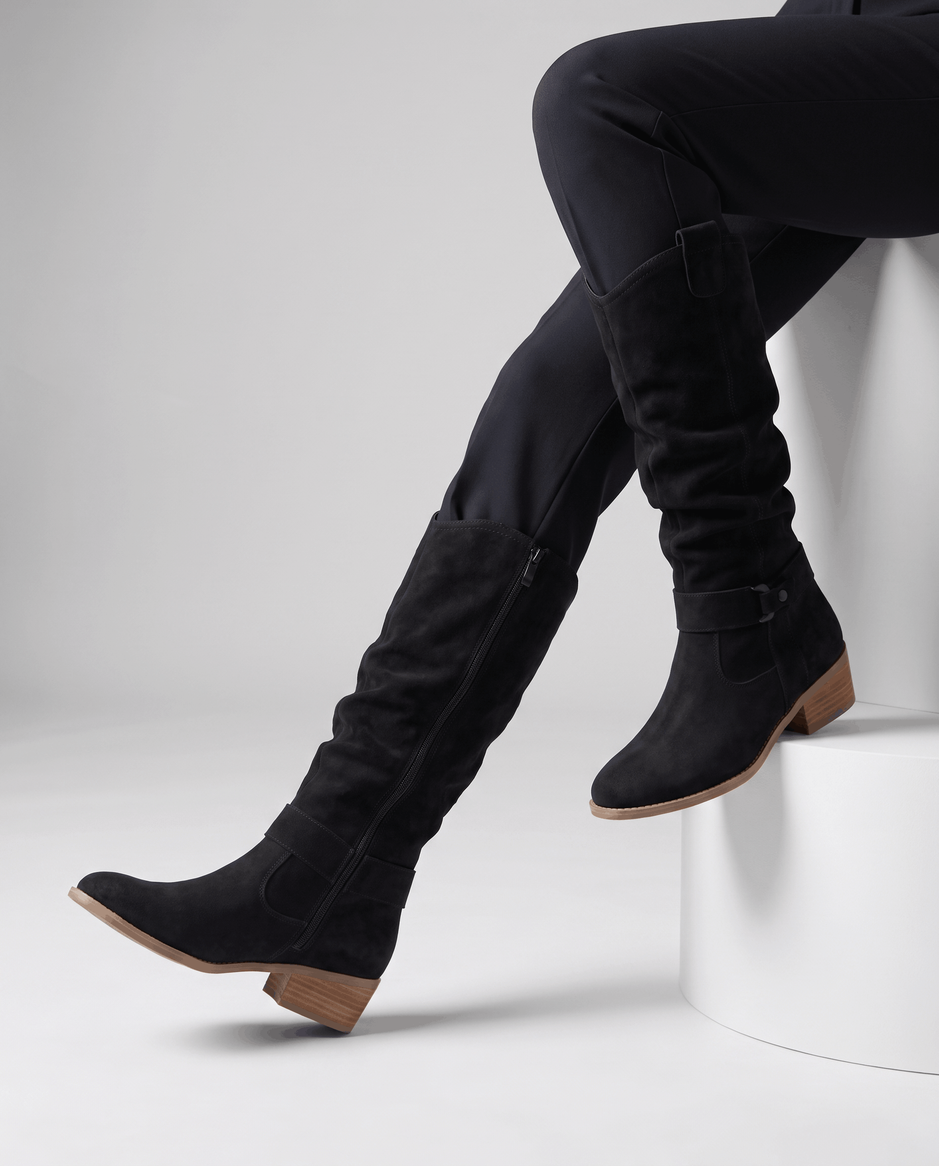 Woman seated wearing black vegan suede knee-high slouch boots with black trousers.