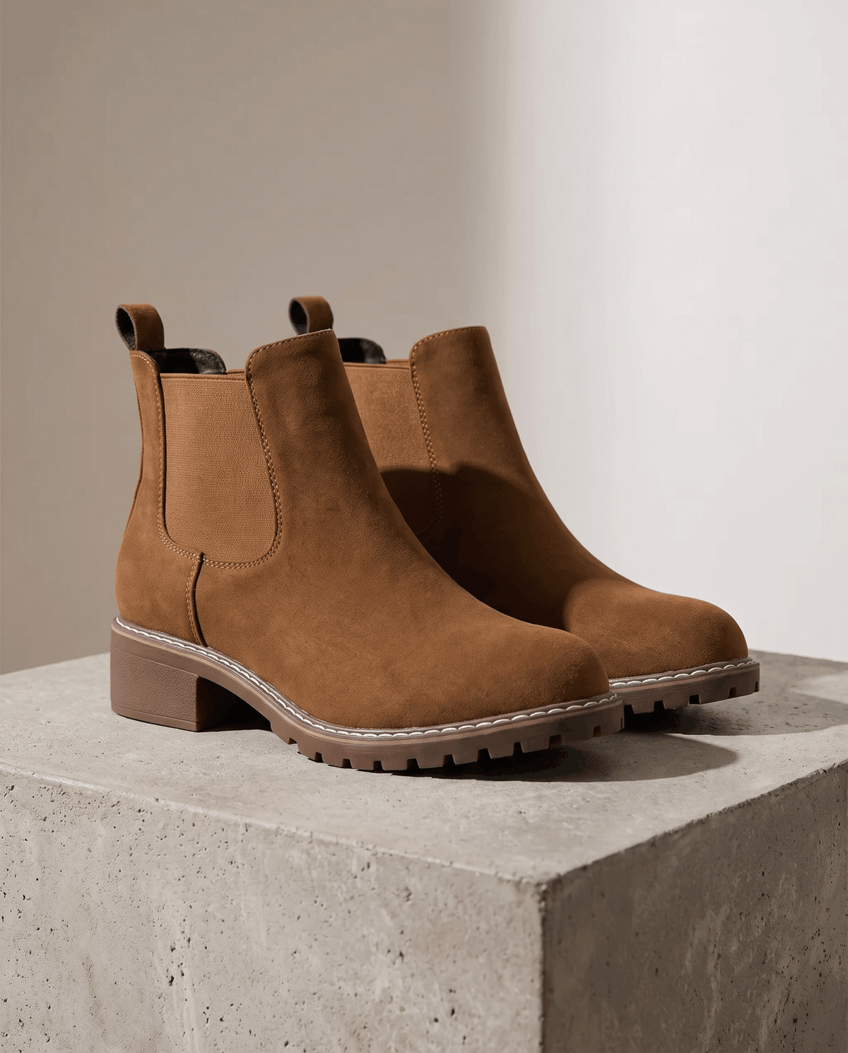 Brown vegan suede Chelsea boots with elastic side panels, a stitched welt, and a chunky lug outsole on a concrete block.