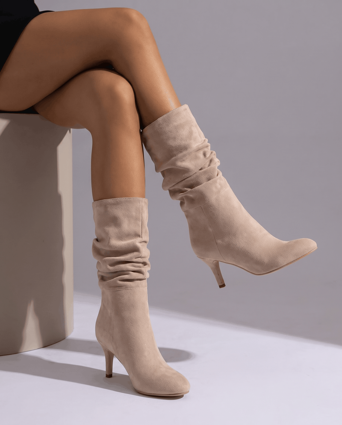 Light beige vegan suede mid-calf boots with a slouched shaft, almond toe, and slim flared heel worn on a seated model.