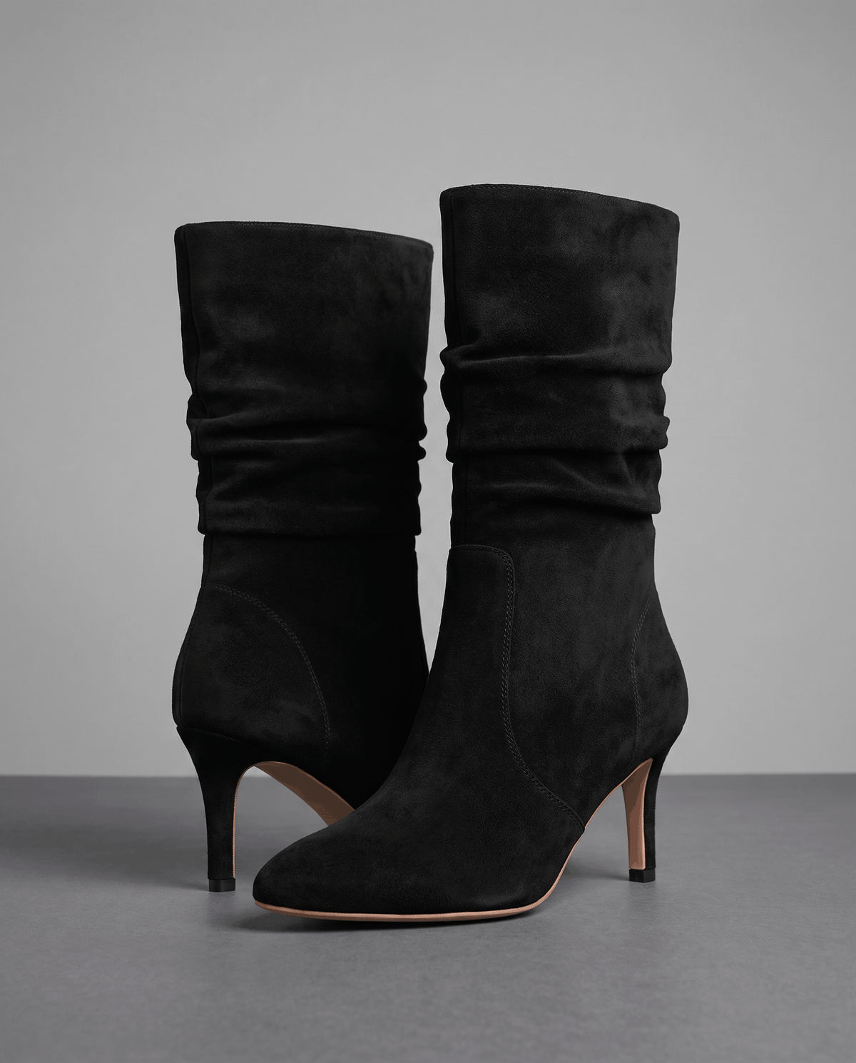 Black vegan suede mid-calf boots with a slouchy shaft, almond toe, and slim flared heel on a grey background.