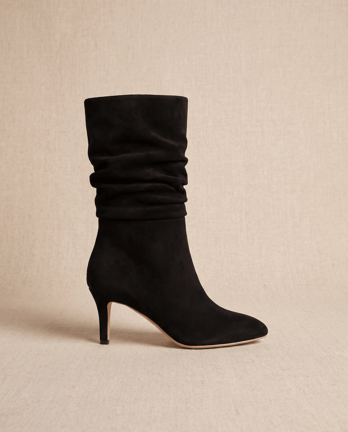 Black vegan suede mid-calf boots with a slouched shaft, almond toe, and slim flared heel on a neutral textured background.