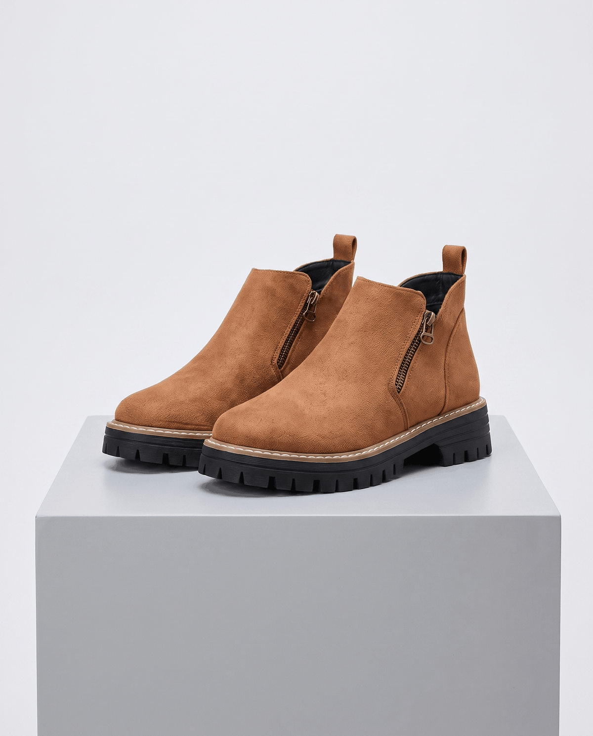 Tan vegan suede ankle boots with side zips, a chunky black lug platform, and cushioned wedge sole on a grey pedestal.