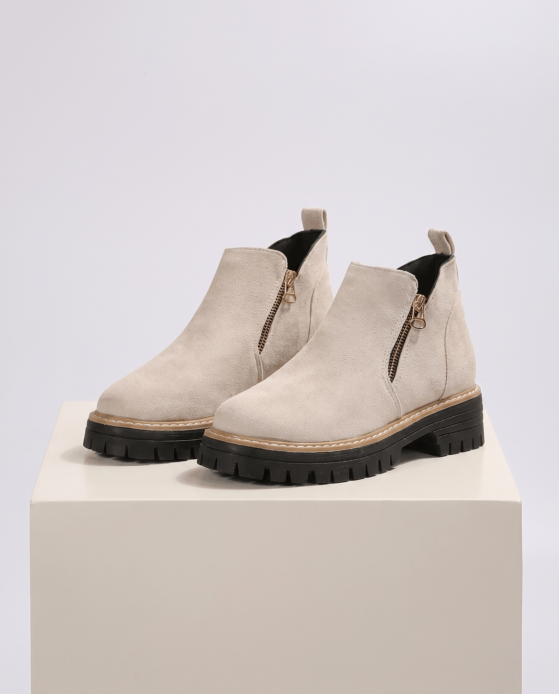 Beige vegan suede ankle boots with side zips and chunky black lug platform wedge soles on a neutral pedestal.