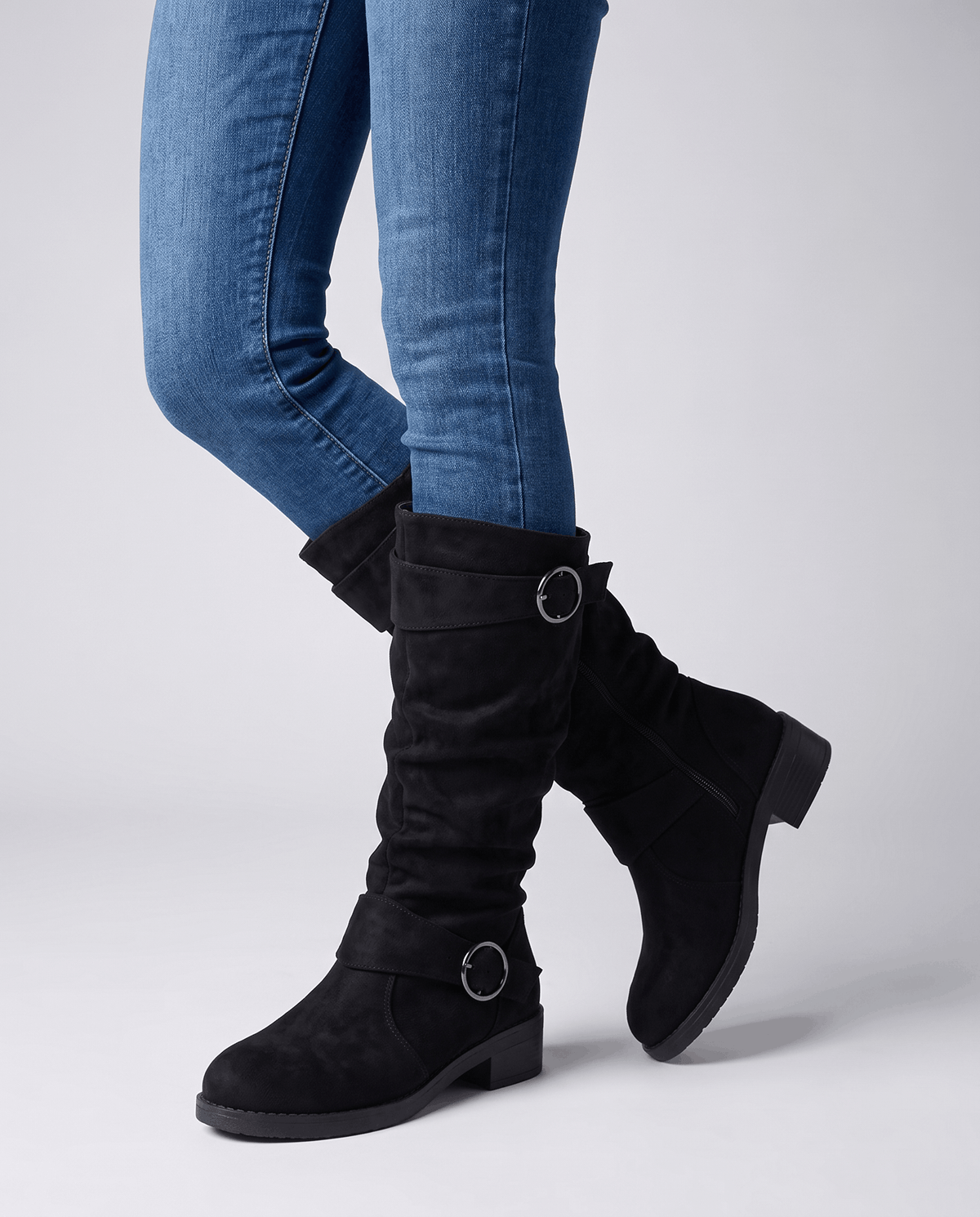 Black vegan suede knee-high boots with a slouched shaft, twin ring straps, and a low stacked heel worn over blue jeans.