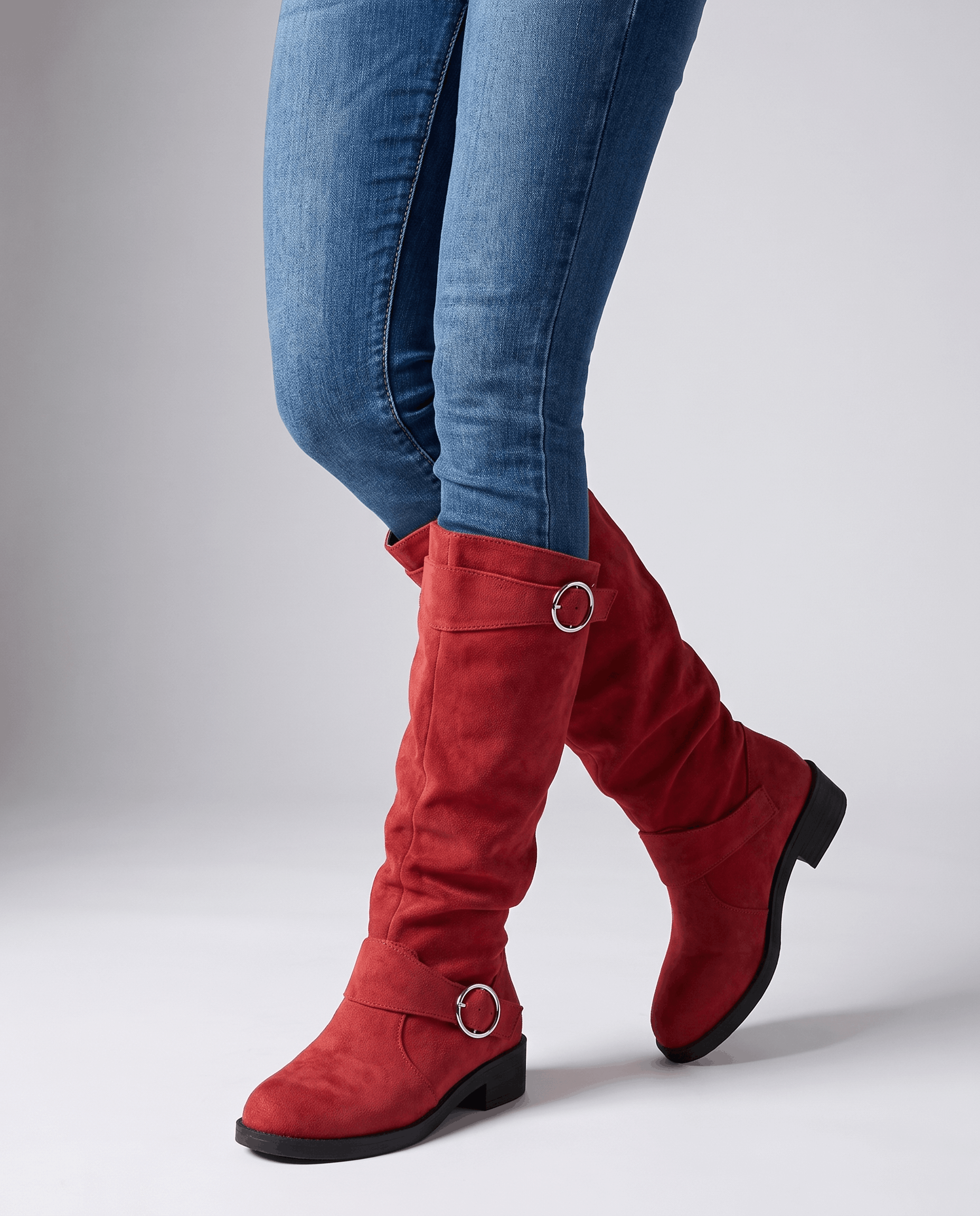 Red vegan suede knee-high boots with a slouched shaft, twin ring straps, and a low stacked heel worn over blue jeans.