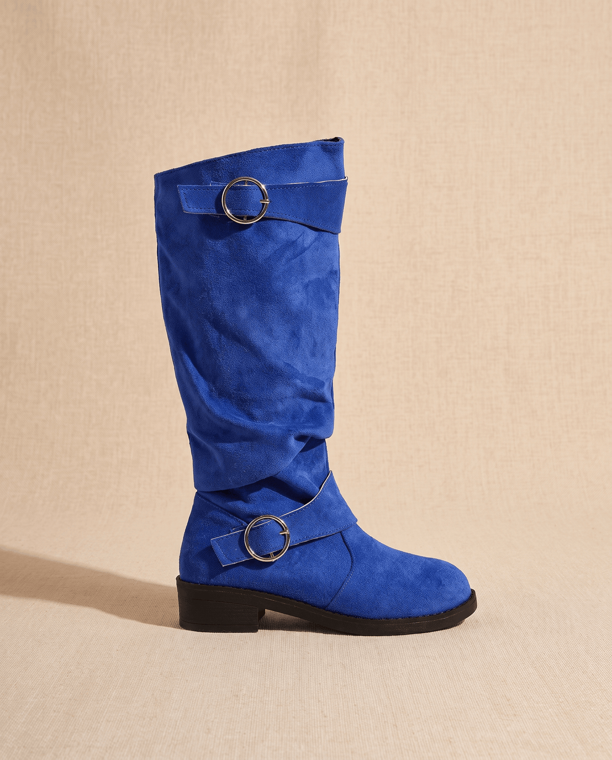 Blue vegan suede knee-high boots with a slouched shaft, round toe, low stacked heel, and twin decorative ring straps.