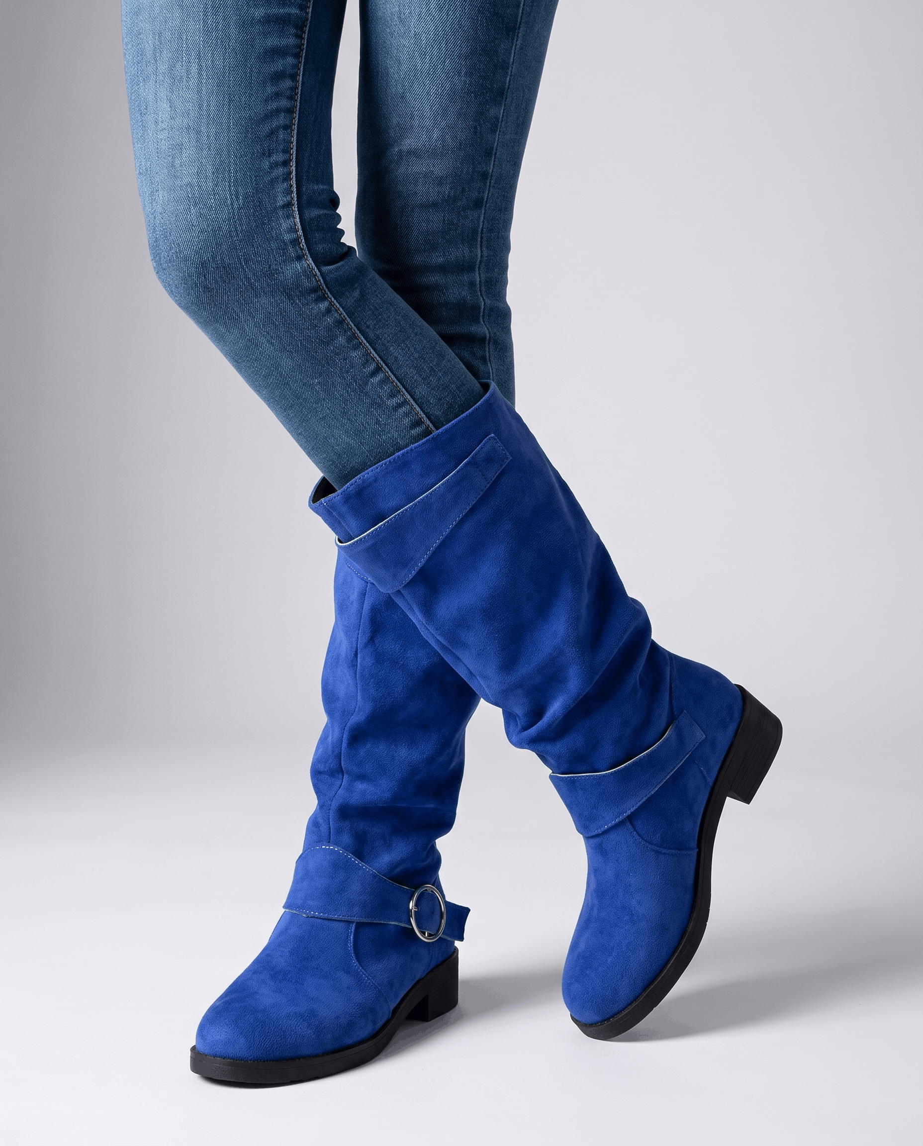 Blue vegan suede knee-high boots with a slouched shaft, twin ring straps, and a low stacked heel worn over blue jeans.