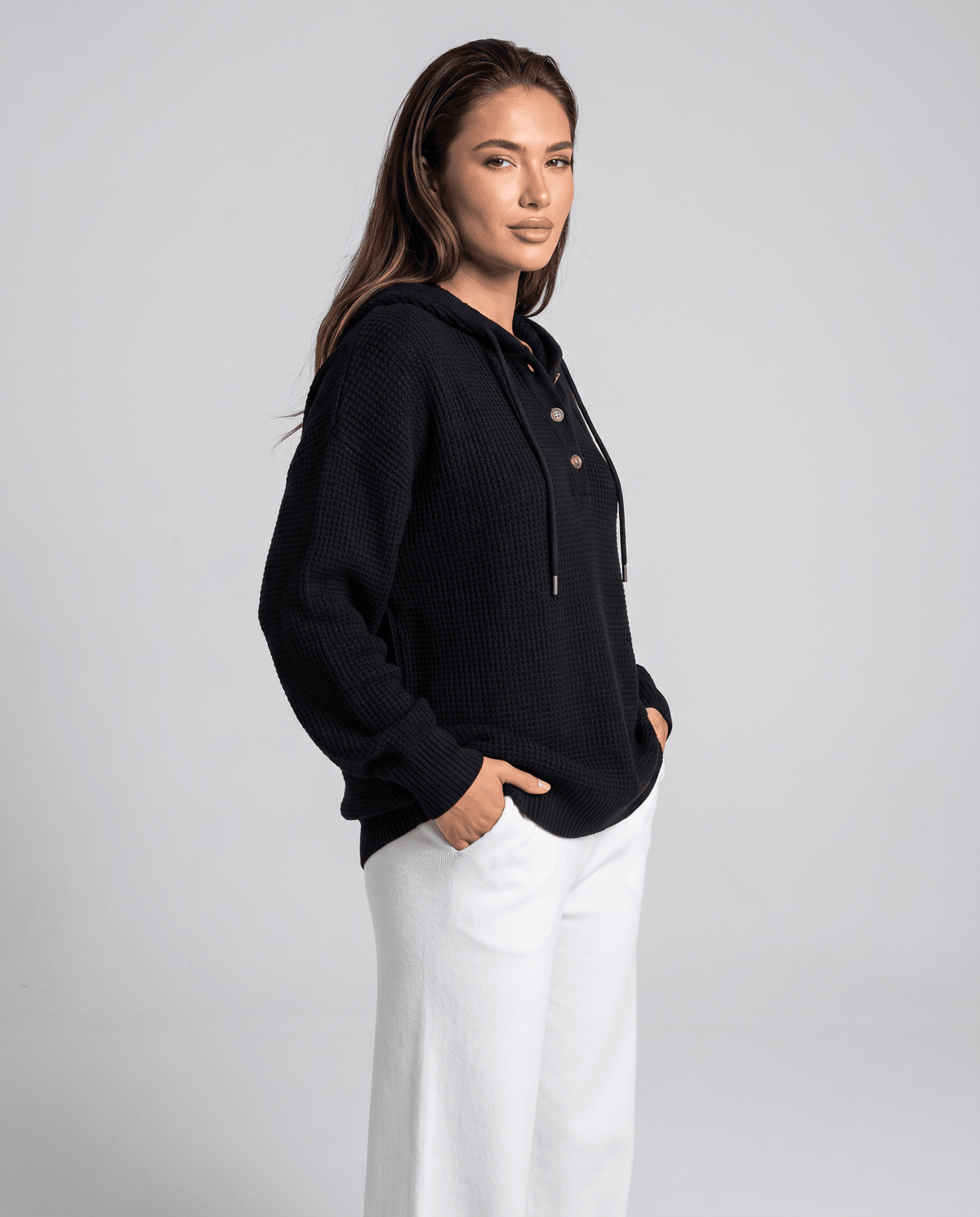 Side angle of the black knit hoodie with hands in pockets and white pants