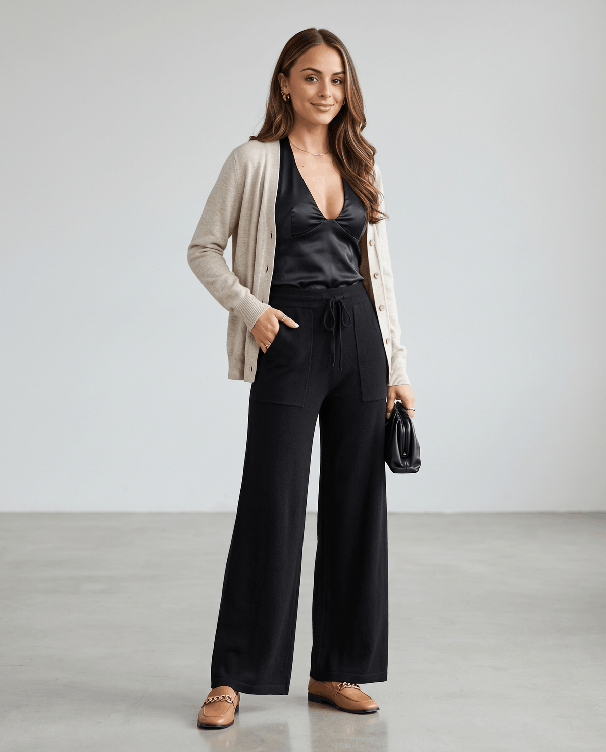 Model in beige cardigan and black satin top wearing black knit pants